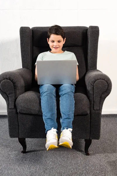 Menino sorridente em jeans sentado em poltrona e usando laptop — Fotografia de Stock