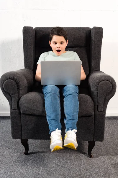 Chocado chico en jeans sentado en sillón y el uso de ordenador portátil - foto de stock