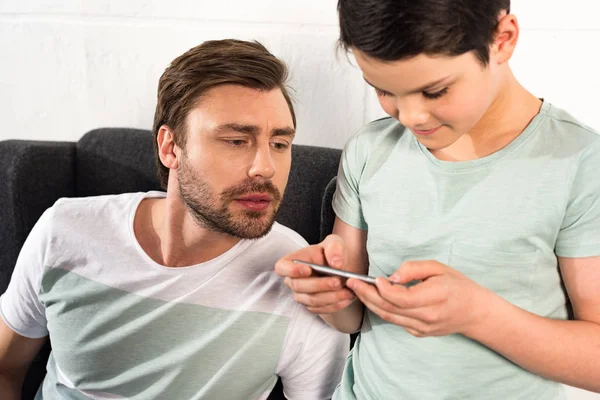 Любопытный отец наблюдает за сыном с помощью смартфона в гостиной — стоковое фото