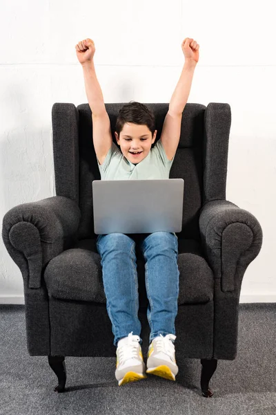 Excited boy in jeans sitting in armchair and using laptop — Stock Photo