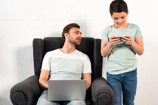 Hijo usando teléfono inteligente y padre usando el ordenador portátil en la sala de estar - foto de stock