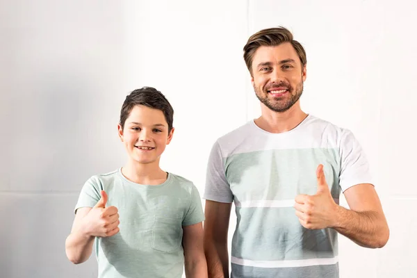 Vista frontal de sonriente hijo y padre mostrando los pulgares hacia arriba en casa - foto de stock