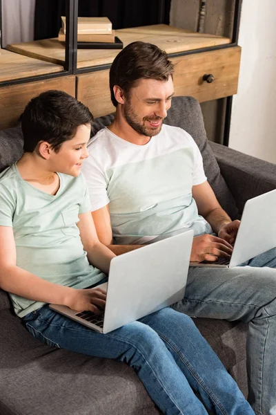 Sonriente hijo y papá usando computadoras portátiles en la sala de estar - foto de stock