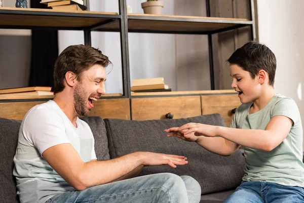 Laughing son and dad sitting on sofa and having fun in living room — Stock Photo