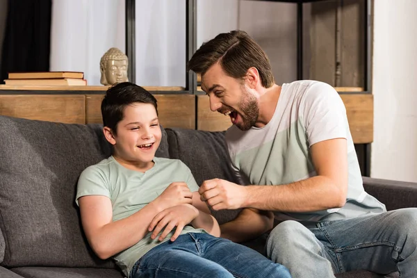 Laughing son and dad sitting on sofa and having fun in living room — Stock Photo