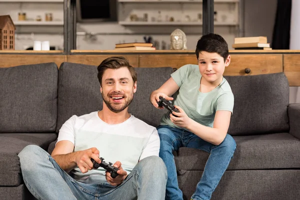 KYIV, UCRANIA - 17 DE ABRIL DE 2019: padre e hijo sonrientes sosteniendo mandos y jugando videojuegos en la sala de estar - foto de stock