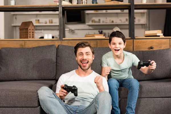 KYIV, UCRANIA - 17 DE ABRIL DE 2019: padre e hijo sonrientes sosteniendo mandos y jugando videojuegos en la sala de estar - foto de stock