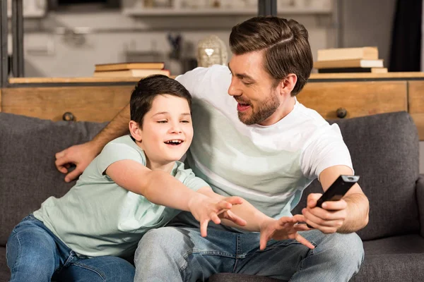 Sonriente hijo y padre con mando a distancia viendo la televisión en la sala de estar - foto de stock