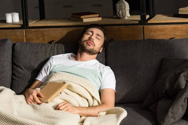 Uomo barbuto con libro dormire sul divano sotto coperta in soggiorno — Foto stock