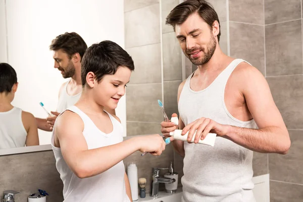Filho e pai aplicando pasta de dentes de manhã no banheiro — Fotografia de Stock