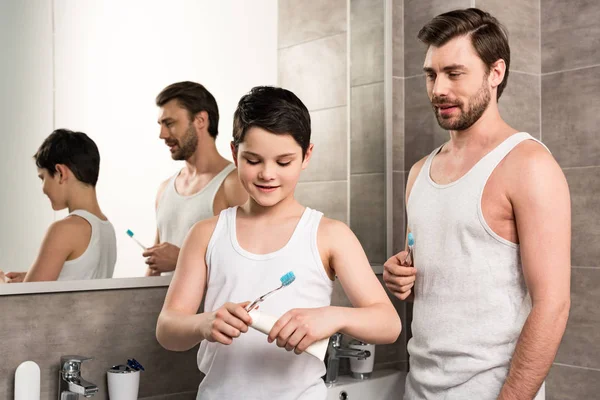 Fils et papa souriants avec brosses à dents et dentifrice dans la salle de bain — Photo de stock