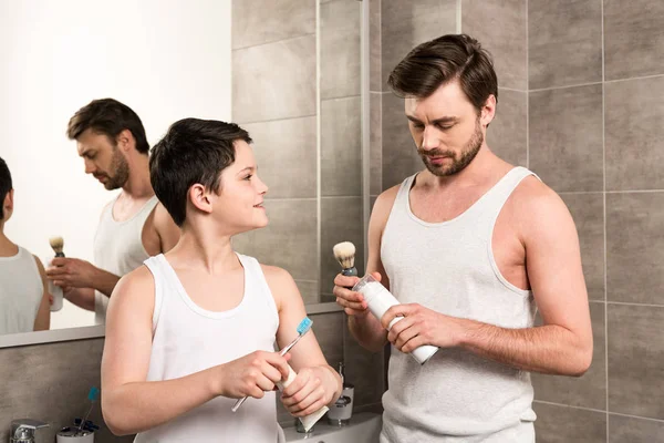 Son brushing teeth and dad shaving in morning in bathroom — Stock Photo