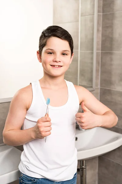 Sourire garçon tenant brosse à dents et montrant pouce dans la salle de bain — Photo de stock