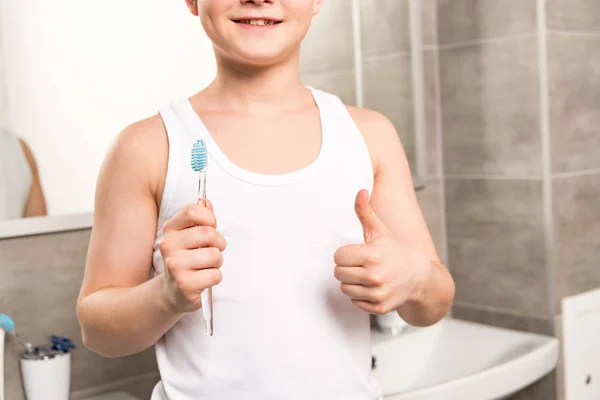 Partial view of smiling boy holding toothbrush and showing thumb up in bathroom — Stock Photo