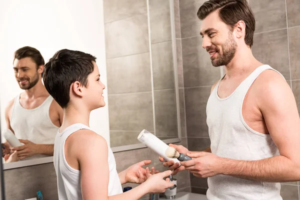 Sorridente papà che dà schiuma da barba e rasoio da barba a figlio in bagno — Foto stock