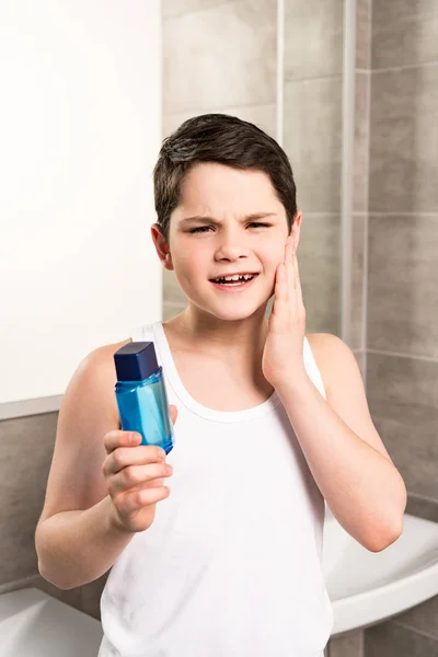 Insatisfait préadolescent garçon appliquant lotion sur le visage et en regardant caméra dans la salle de bain — Photo de stock