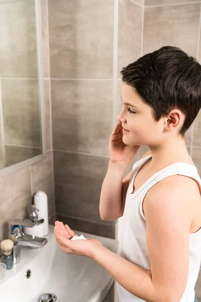 Sourire garçon appliquant mousse à raser sur le visage dans la salle de bain — Photo de stock