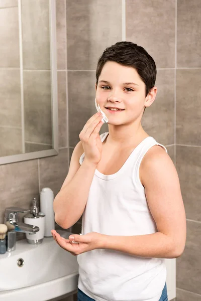 Smiling boy applying shaving foam on face in bathroom — Stock Photo