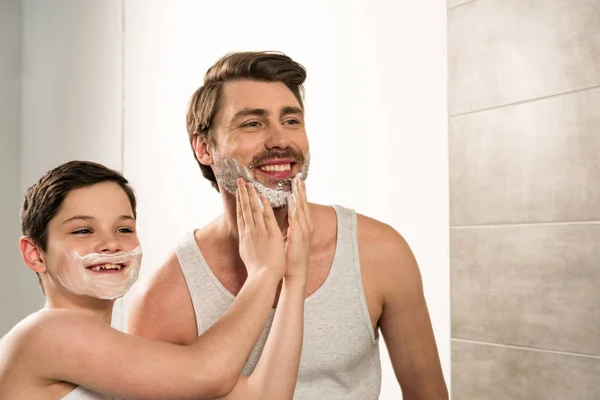 Preteen boy applying shaving foam on smiling father in bathroom — Stock Photo