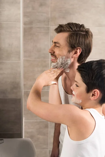 Niño preadolescente aplicando espuma de afeitar en el padre sonriente en el baño - foto de stock