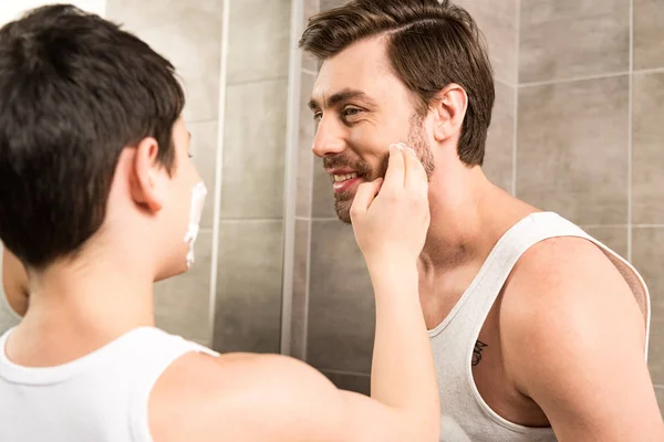 Niño preadolescente aplicando espuma de afeitar en el padre sonriente en el baño - foto de stock