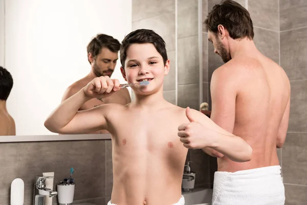Сын чистить зубы рядом с отцом и показывая большой палец вверх в ванной комнате — стоковое фото
