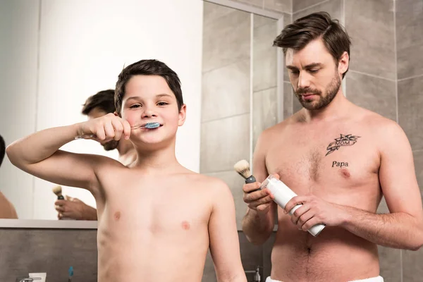 Preteen menino escovar dentes e pai segurando espuma de barbear e escova no banheiro — Fotografia de Stock