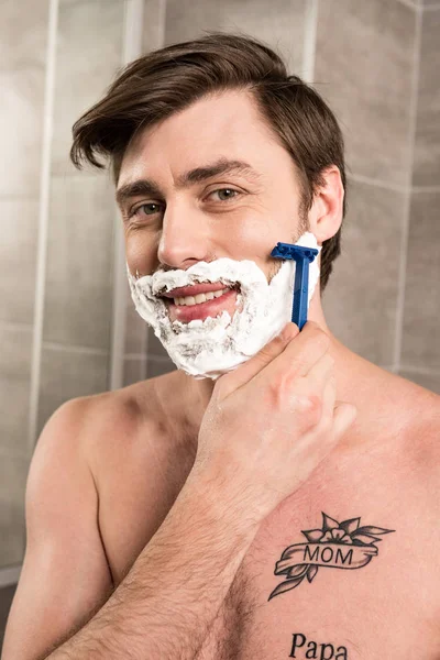 Улыбающийся мужчина бреет бороду с бритвой в ванной комнате — стоковое фото