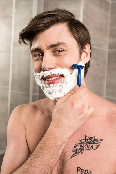 Lächelnder Mann rasiert Bart mit Rasiermesser im Badezimmer — Stockfoto