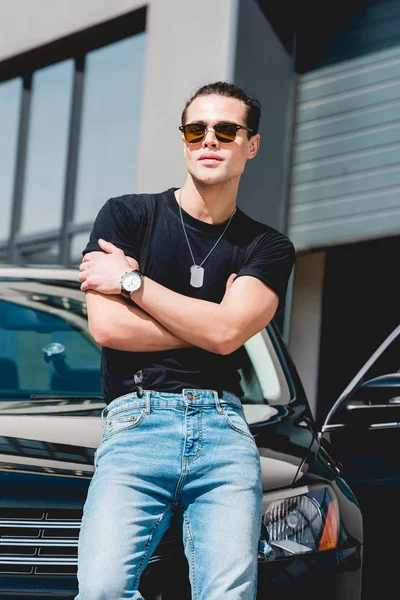 Bello uomo elegante in occhiali da sole in posa vicino auto con braccia incrociate — Foto stock