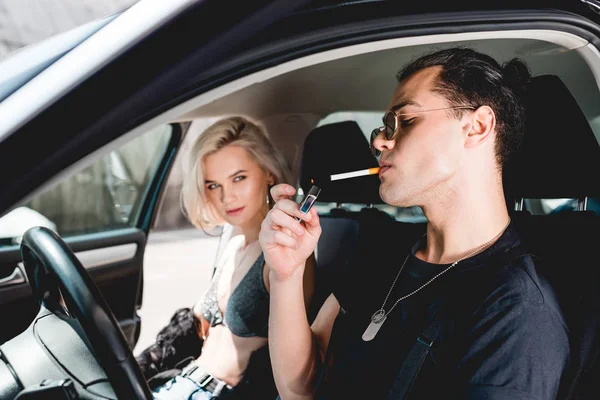 Stylish handsome man lighting cigarette while sitting with beautiful girl in car — Stock Photo