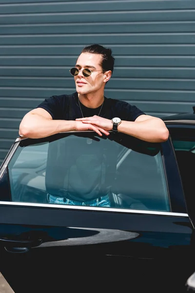Guapo hombre con estilo en gafas de sol posando cerca de coche y mirando hacia otro lado - foto de stock
