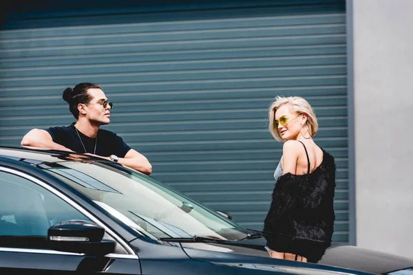 Elegante hombre guapo y hermosa chica en gafas de sol posando cerca de coche - foto de stock