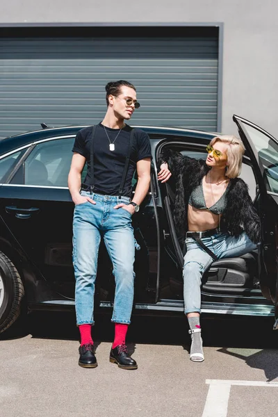 Elegante hombre guapo y hermosa chica en gafas de sol posando cerca de coche - foto de stock