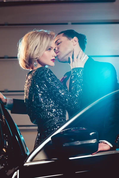 Stylish man kissing beautiful seductive woman near car with blue light — Stock Photo