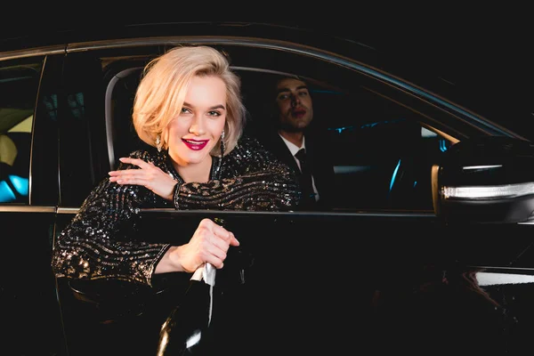 Stilvoller Mann und schöne lächelnde junge Frau in formeller Kleidung mit Champagnerflasche im Auto sitzend — Stockfoto