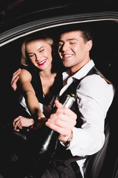 Stylish happy man and woman with champagne bottle in car — Stock Photo