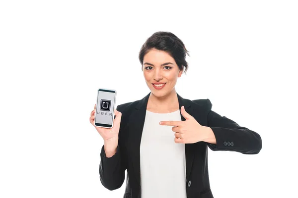 KYIV, UCRANIA - 24 DE ABRIL DE 2019: Hermosa mujer de negocios de raza mixta que señala con el dedo al teléfono inteligente con la aplicación Uber en la pantalla mientras sonríe a la cámara aislada en blanco . - foto de stock