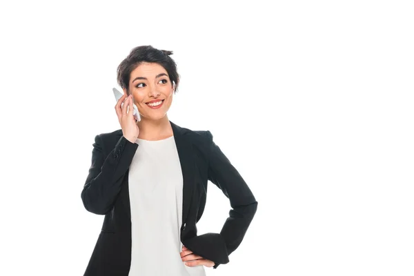 Sonriente mujer de negocios de raza mixta hablando en el teléfono inteligente y sosteniendo la mano en la cadera aislado en blanco - foto de stock