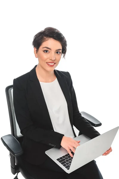 Sonriente mujer de negocios de raza mixta utilizando el ordenador portátil mientras está sentado en la silla de oficina y mirando a la cámara aislada en blanco - foto de stock