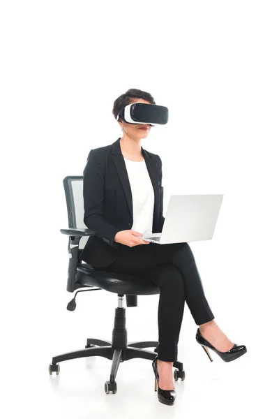 Young mixed race businesswoman in virtual reality headset using laptop while sitting in office chair on white background — Stock Photo