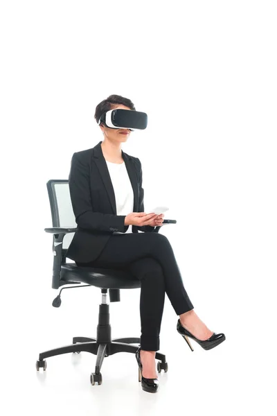 Young mixed race businesswoman in virtual reality headset using laptop while sitting in office chair on white background — Stock Photo