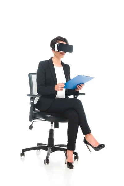 Femme d'affaires mixte utilisant casque de réalité virtuelle tout en étant assis dans la chaise de bureau et tenant presse-papiers sur fond blanc — Photo de stock
