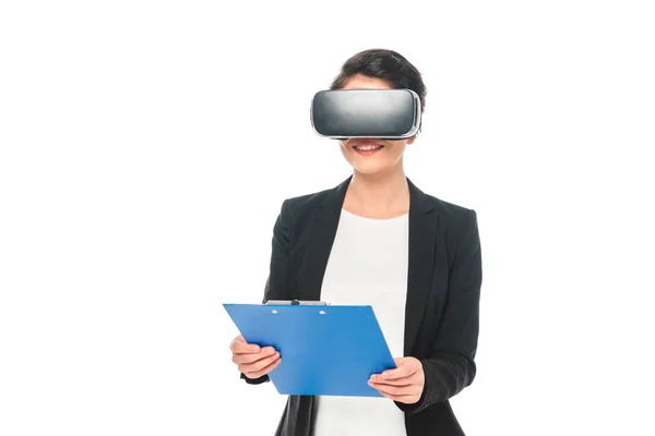 Sonriente mujer de negocios de raza mixta usando auriculares de realidad virtual y sujetando el portapapeles aislado en blanco - foto de stock