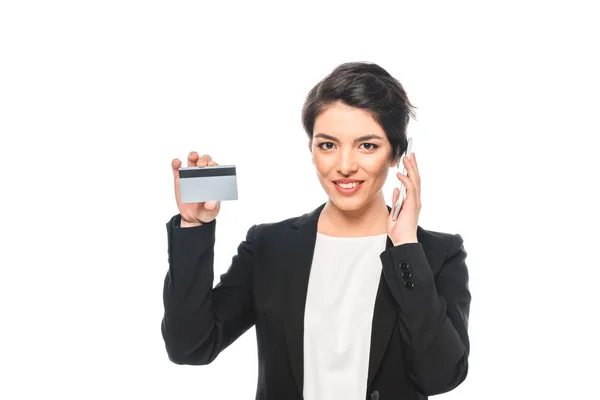 Alegre mixta mujer de negocios que muestra la tarjeta de crédito mientras habla en el teléfono inteligente aislado en blanco - foto de stock