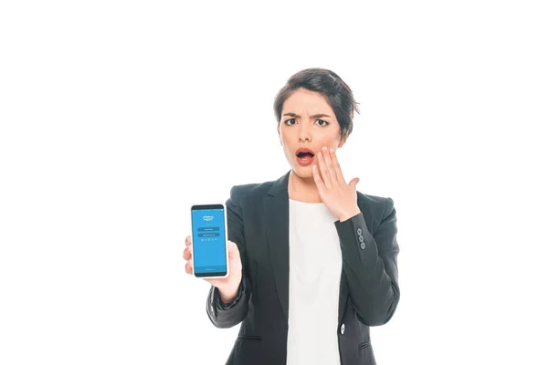 KYIV, UCRAINA - 24 APRILE 2019: Scioccata donna d'affari mista che mostra smartphone con app Skype sullo schermo isolato su bianco . — Foto stock