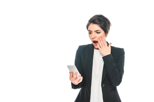 Sorprendida mujer de negocios de raza mixta usando smartphone y sosteniendo la mano cerca de la cara aislada en blanco - foto de stock