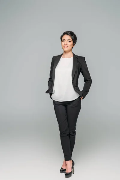 Lächelnde gemischte Geschäftsfrau mit Händen in den Taschen und Blick in die Kamera auf grauem Hintergrund — Stockfoto