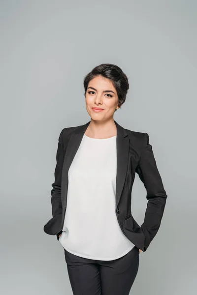 Bastante mixta mujer de negocios de pie con las manos en el bolsillo y sonriendo a la cámara aislada en gris - foto de stock