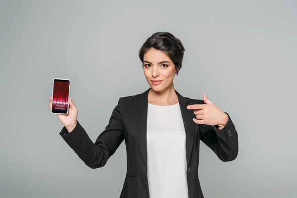 Beautiful mixed race businesswoman showing smartphone with trading courses app on screen isolated on grey — Stock Photo